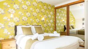 Une chambre d’hôtel avec un papier peint à motifs floraux, un grand lit avec une literie blanche, une table de chevet en bois et un miroir.