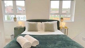 A neatly made bed with a green blanket and two rolled white towels, two bedside lamps, a small green nightstand, and a window with a view of buildings.