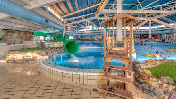An indoor swimming pool with a slide, a wooden play structure, and a thatched-roof pavilion.
