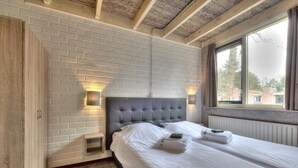 A hotel room with a brick-patterned wall, a tufted headboard, a large window with curtains, and neatly made beds.