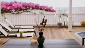 A wooden deck with lounge chairs, a table with a vase and a bottle, and a white fence with pink flowers in the background. - T3 Pisicine Chauffée et Privative (Saint-Leu)