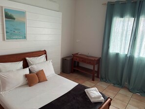 A bedroom with a bed, pillows, a nightstand, a desk, a trash bin, and a window with curtains.