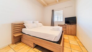 A modern bedroom with a wooden bed, a TV on a wooden cabinet, and a window with blinds. - T3 Pisicine Chauffée et Privative (Saint-Leu)