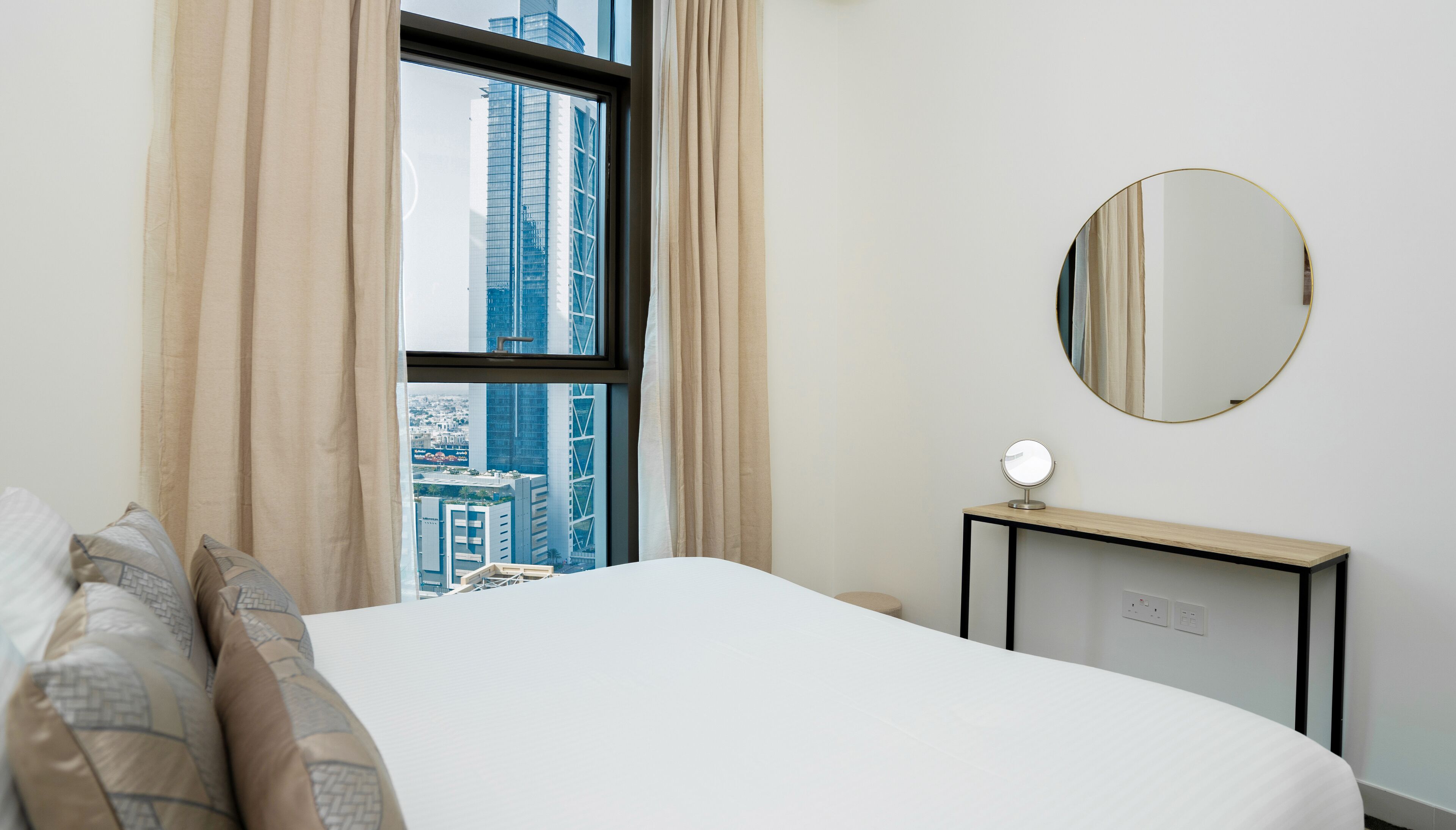 A hotel room with a bed, a bedside table with a mirror, and a view of tall buildings through a window with beige curtains.