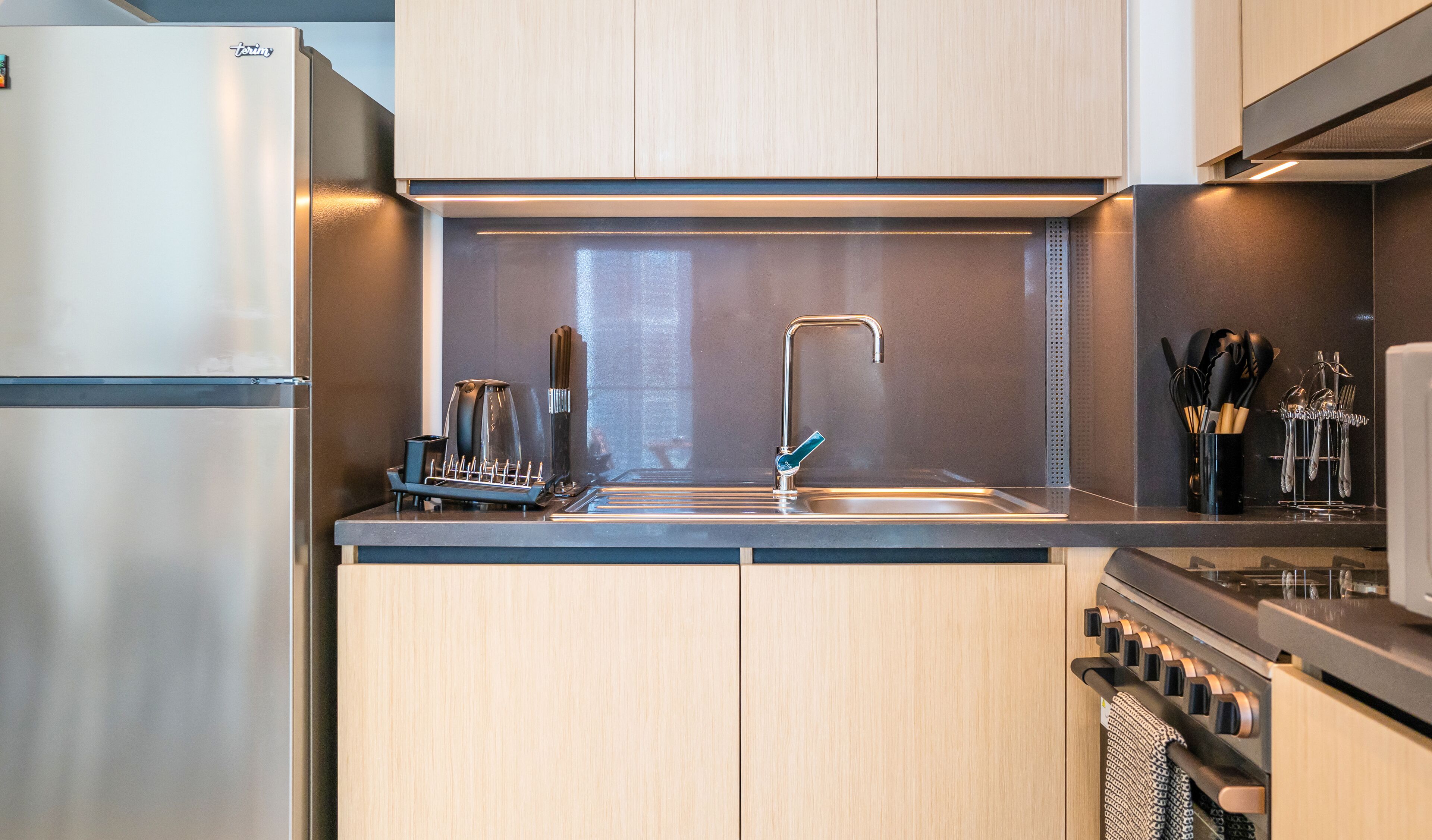 A modern kitchen with a stainless steel refrigerator, a built-in oven, a sink with a faucet, and a coffee maker.