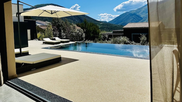 Un área de piscina moderna al aire libre con sillones reclinables y una sombrilla, rodeada de montañas y vegetación.