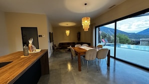 Un comedor moderno con una mesa y sillas de madera, una araña y vistas a un paisaje montañoso a través de amplios ventanales.