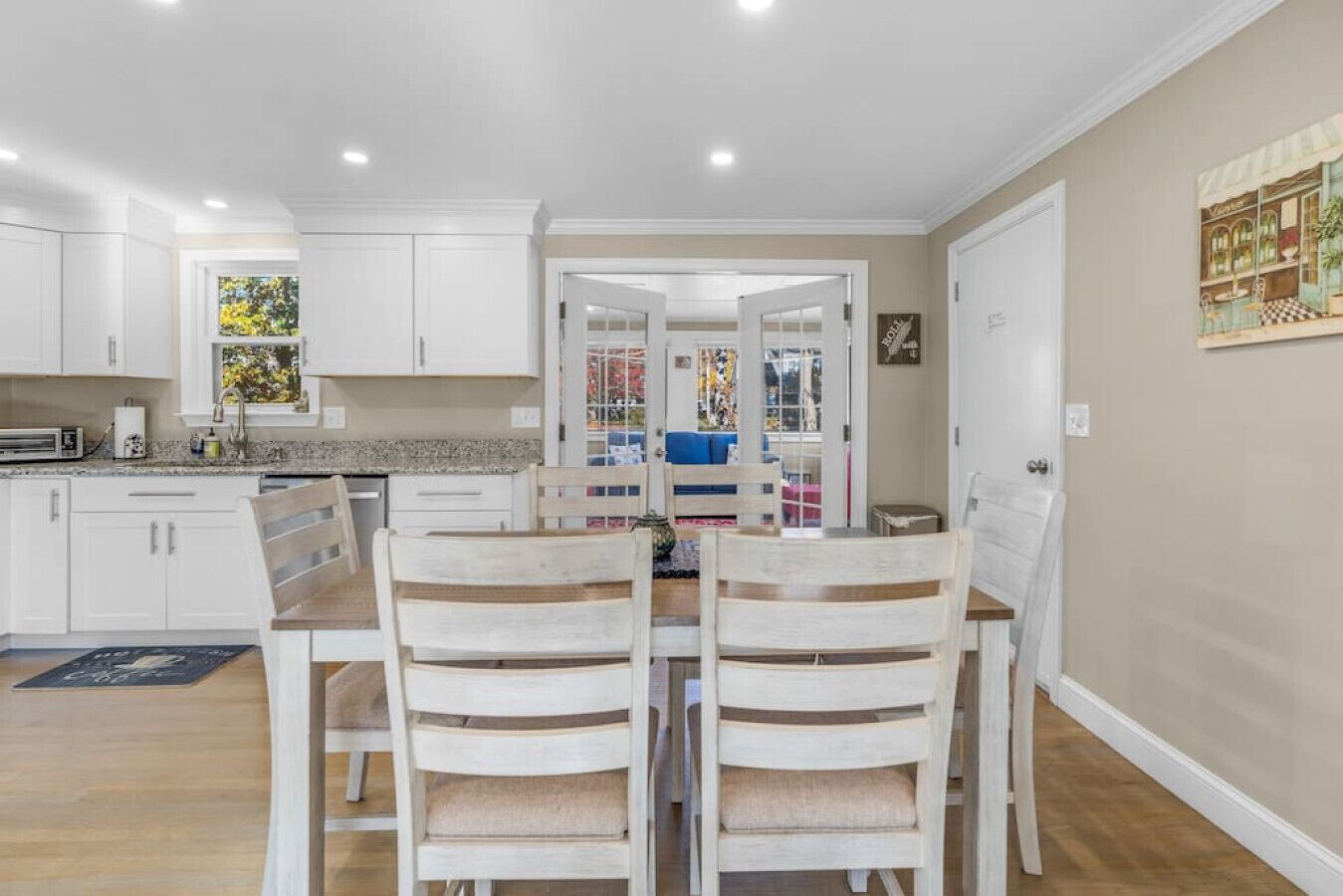 A modern kitchen with white cabinets, a dining area with wooden chairs, and a view into the living room.