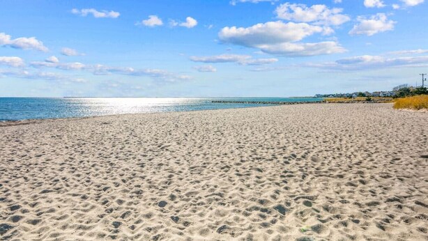 Ein weiter Sandstrand mit Fußspuren, die zum Wasser führen, ein klarer Himmel und Gebäude in der Ferne.