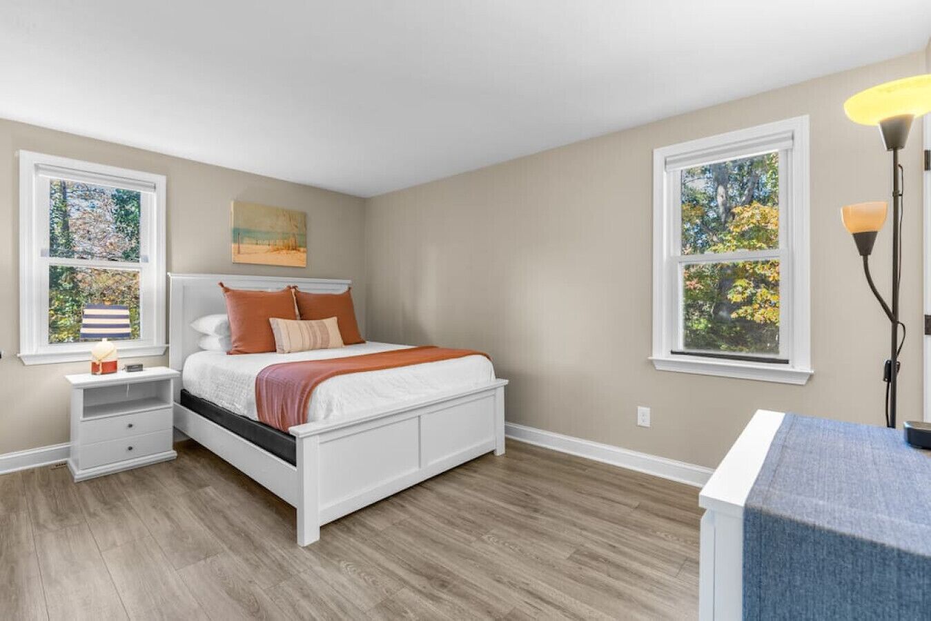 A bedroom with a bed, nightstands, a lamp, and two windows with views of trees.