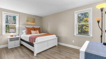 A bedroom with a bed, nightstands, a lamp, and two windows with views of trees.