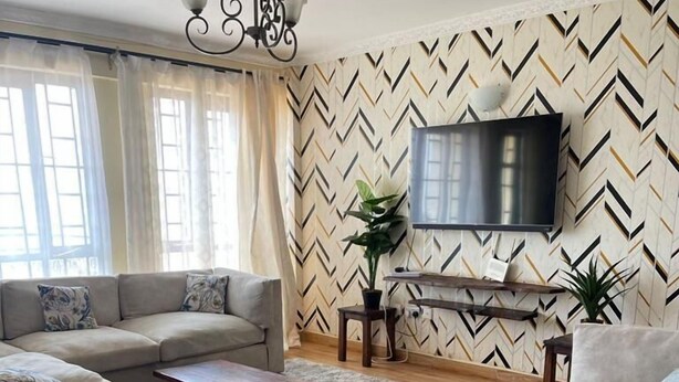 A living room with a sectional sofa, a coffee table, a TV, and a patterned wall. - Ruaka view (Ruaka)