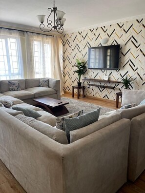 A living room with a sectional sofa, a coffee table, a TV, and a patterned wall. - Ruaka view (Ruaka)