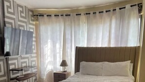 A hotel room with a large bed, a TV, a bedside table with a lamp, and a patterned rug. - Ruaka view (Ruaka)