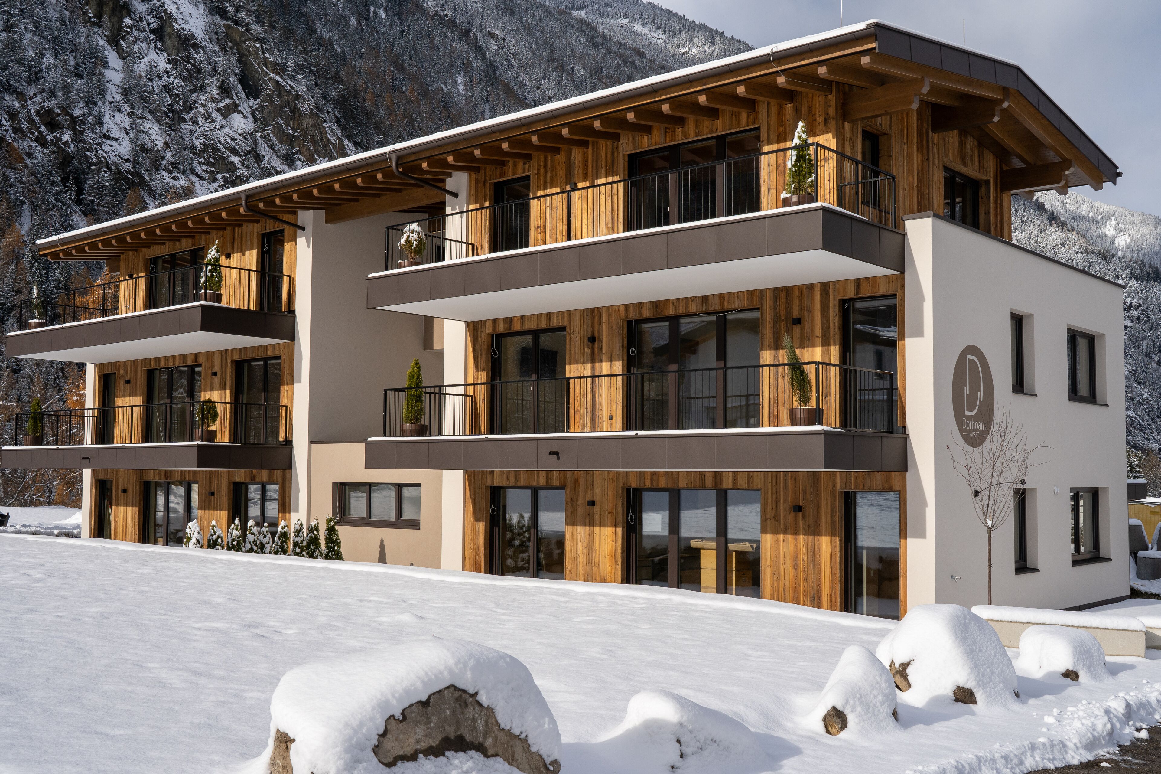 A modern two-story building with wooden exterior and balconies, set against a snowy mountain backdrop.