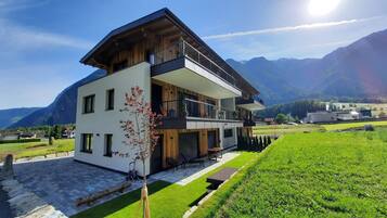 A modern two-story house with a balcony, surrounded by a well-maintained lawn and mountains in the background.