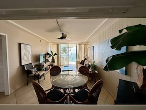 A room with a round glass table, wicker chairs, a ceiling fan, a TV, and a large plant.