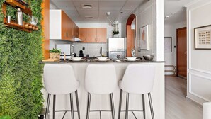 A modern kitchen with a green vertical garden, white bar stools, and a central island. - Silkhaus Manchester Tower, Dubai Marina (Dubai)