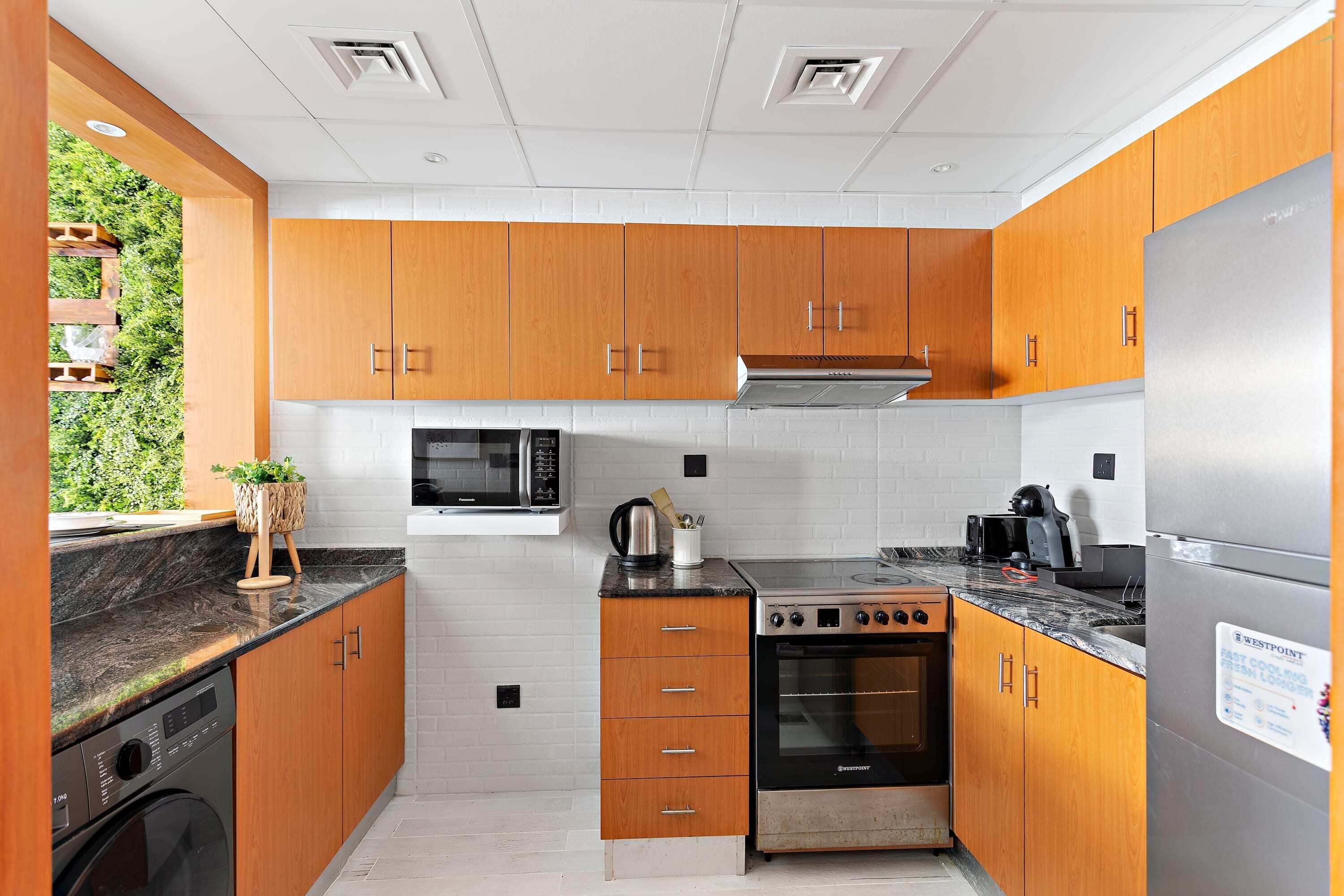 A compact kitchen with wooden cabinets, a stainless steel refrigerator, a microwave, a stove, and a washing machine.