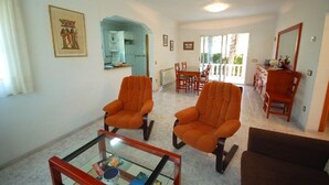 Een ruime woonkamer met twee oranje fauteuils, een bruine bank en een glazen salontafel. Op de achtergrond is een eethoek met houten stoelen en een tafel te zien.