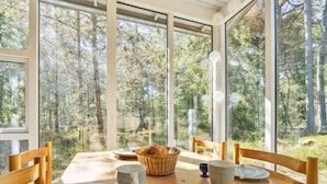 A dining table with chairs, plates, cutlery, a basket of bread, and a cup, set in a room with large windows overlooking a forest.