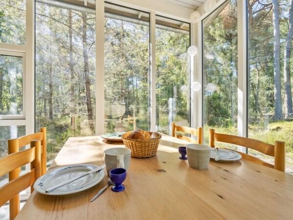 A dining table with chairs, plates, cutlery, a basket of bread, and a cup, set in a room with large windows overlooking a forest.
