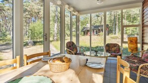 A sunlit room with large windows, wicker chairs, a wooden table with a basket of bread, and a view of a forest and a cabin.