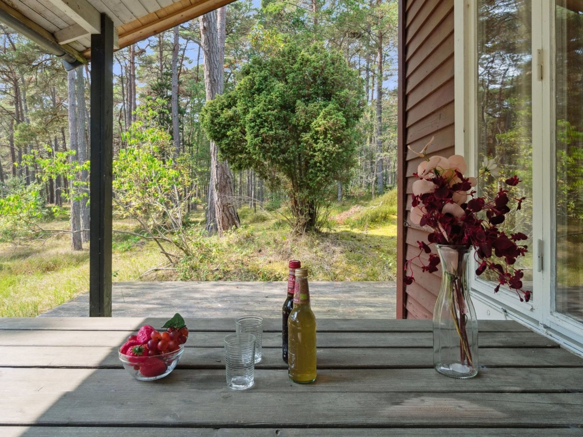 A wooden table with a bowl of fruit, a bottle, and a vase with flowers on a porch overlooking a forest.