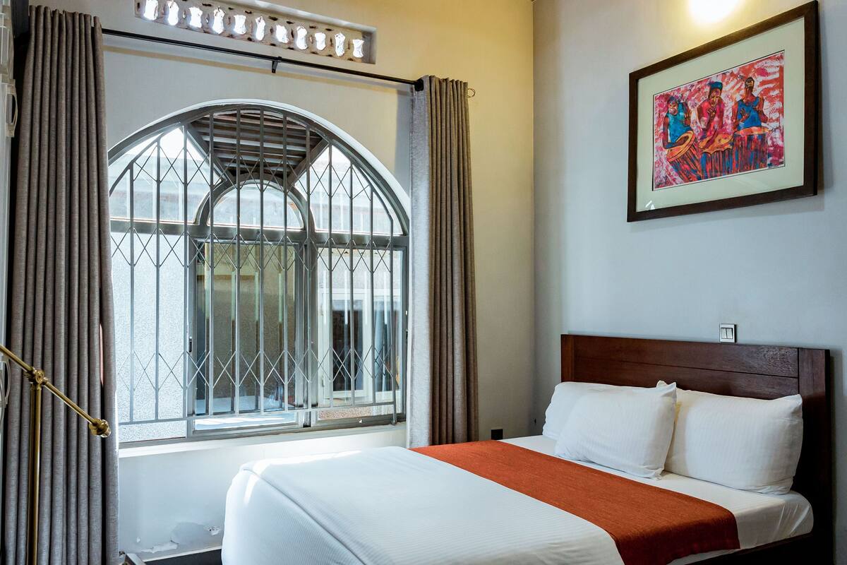 Une chambre d’hôtel dotée d’une grande fenêtre en arc de cercle, d’un lit avec du linge de maison blanc et un plaid orange, ainsi que d’un tableau coloré accroché au mur.