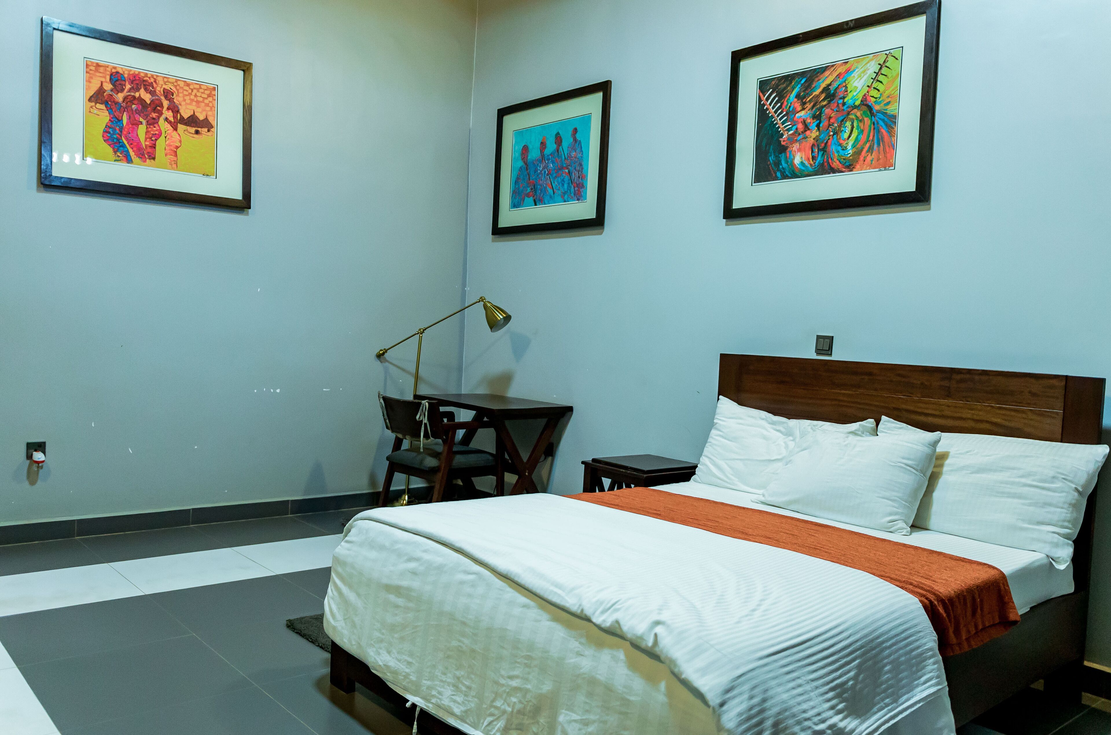 A neatly made bed with white linens and an orange throw blanket, a small wooden side table, and a desk with a lamp. There are three framed artworks on the wall above the bed.