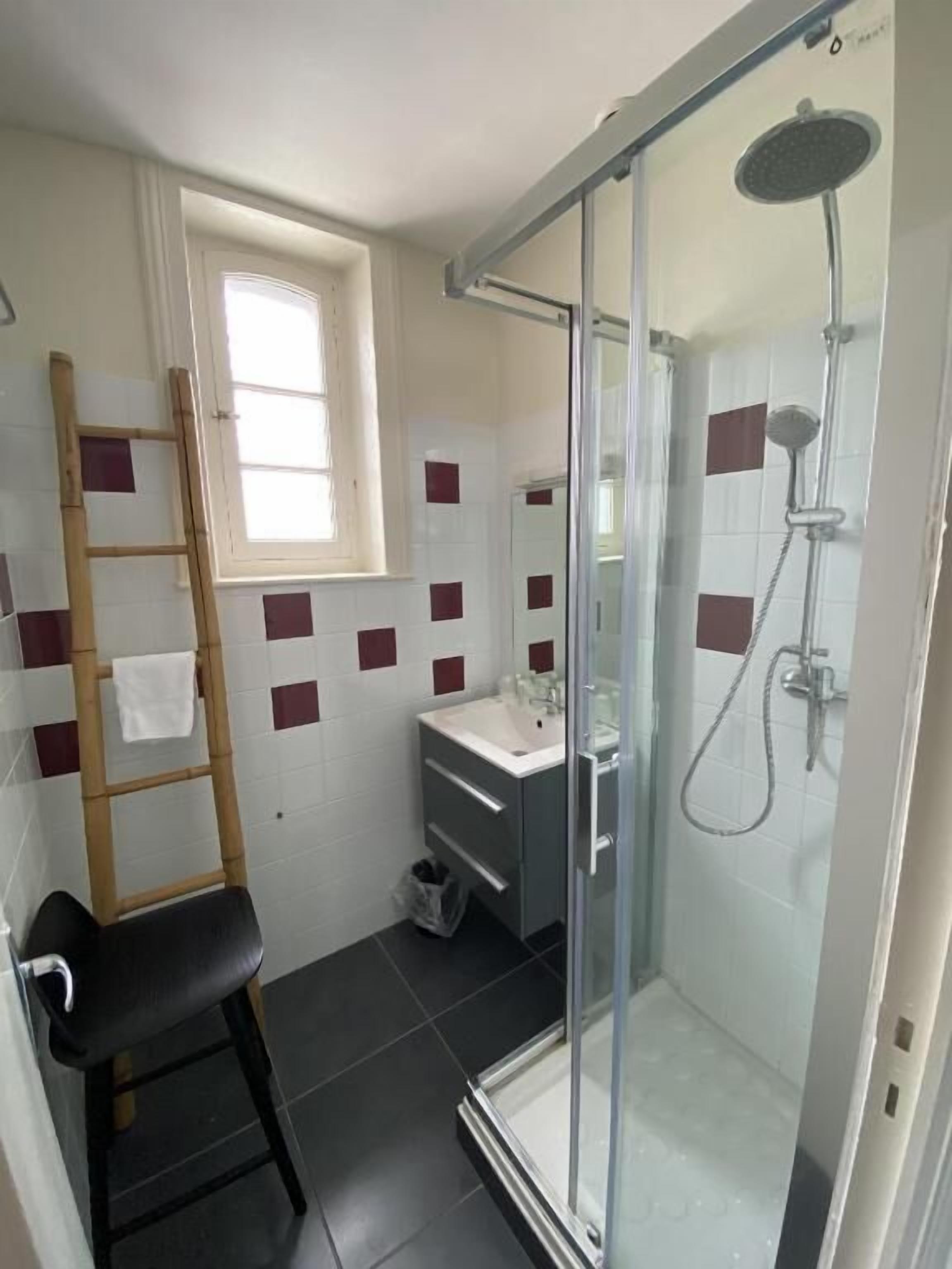 Een badkamer met een glazen douchecabine, een houten ladder, een zwarte stoel en een klein raam.