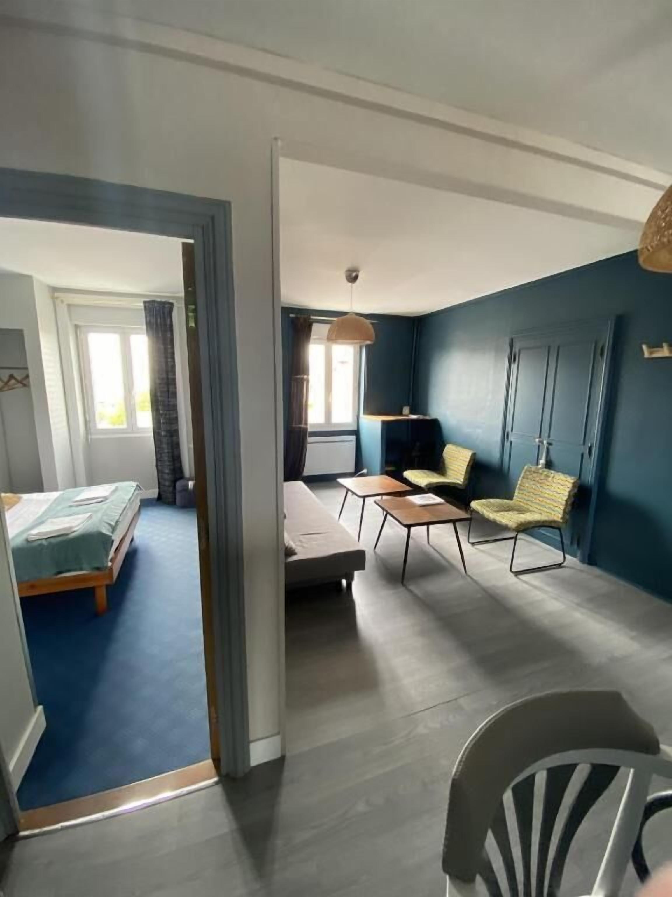A room with a bed, a sofa, a small table, and a chair. There is a window with curtains and a door.