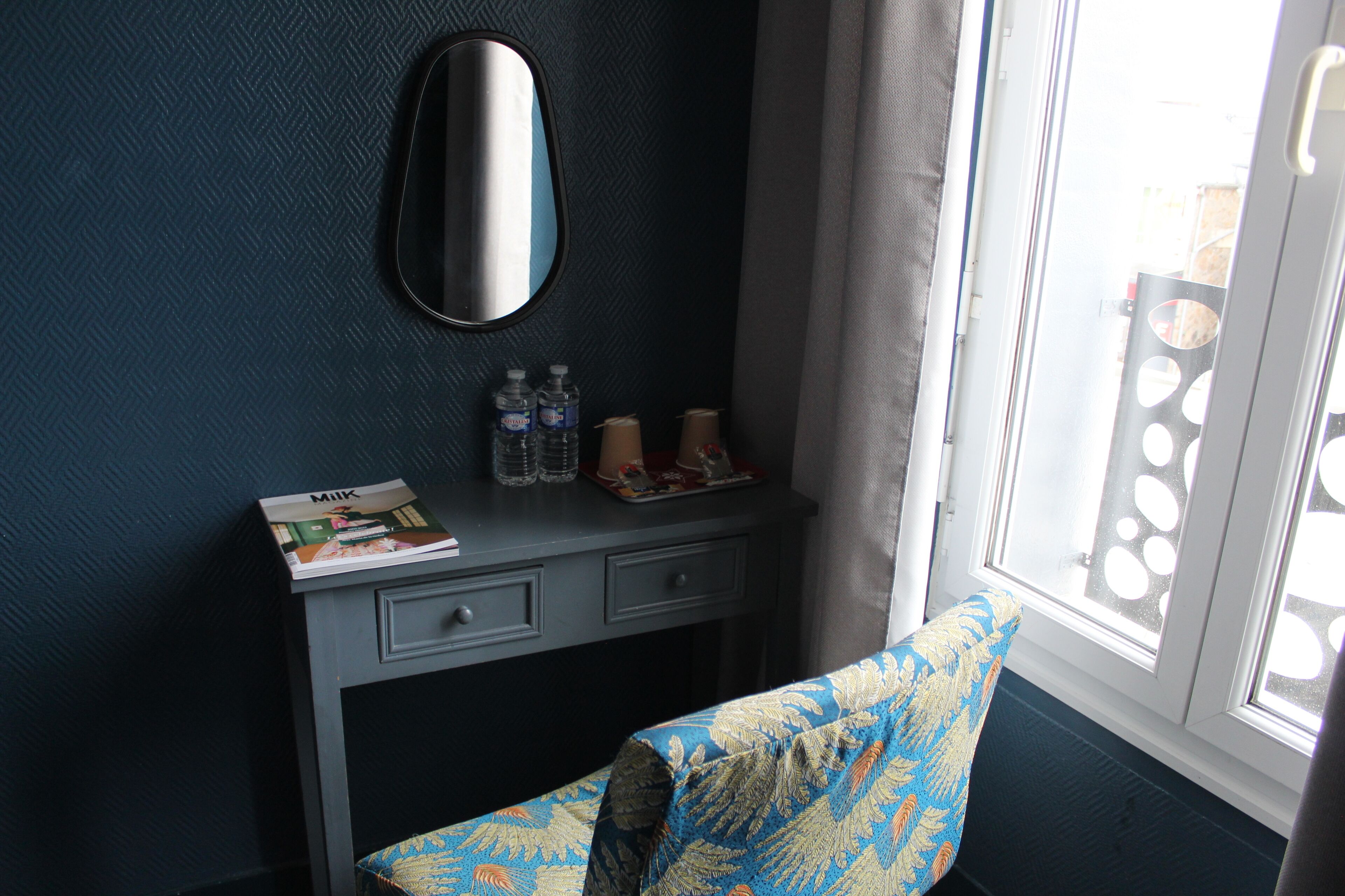 A room with a dark blue wall, a mirror, a small desk with drawers, a chair with a patterned cushion, and a window with a view of the outside.