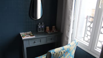 A room with a dark blue wall, a mirror, a small desk with drawers, a chair with a patterned cushion, and a window with a view of the outside.
