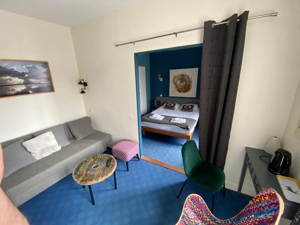 Una habitación con un sofá gris, una mesita redonda pequeña, una silla verde, una silla colorida, una cama con cabecera azul y un cuadro en la pared.