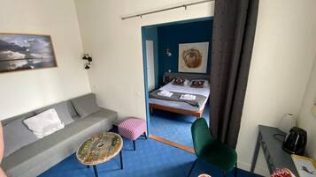 Une pièce avec un canapé gris, une petite table ronde, une chaise verte, une chaise aux couleurs vives, un lit avec une tête de lit bleue et un tableau accroché au mur.