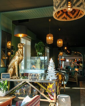 Un interior acogedor con una mesa de madera que exhibe diversos objetos, una estatua dorada de un gato, un árbol de Navidad blanco y lámparas de mimbre colgantes.