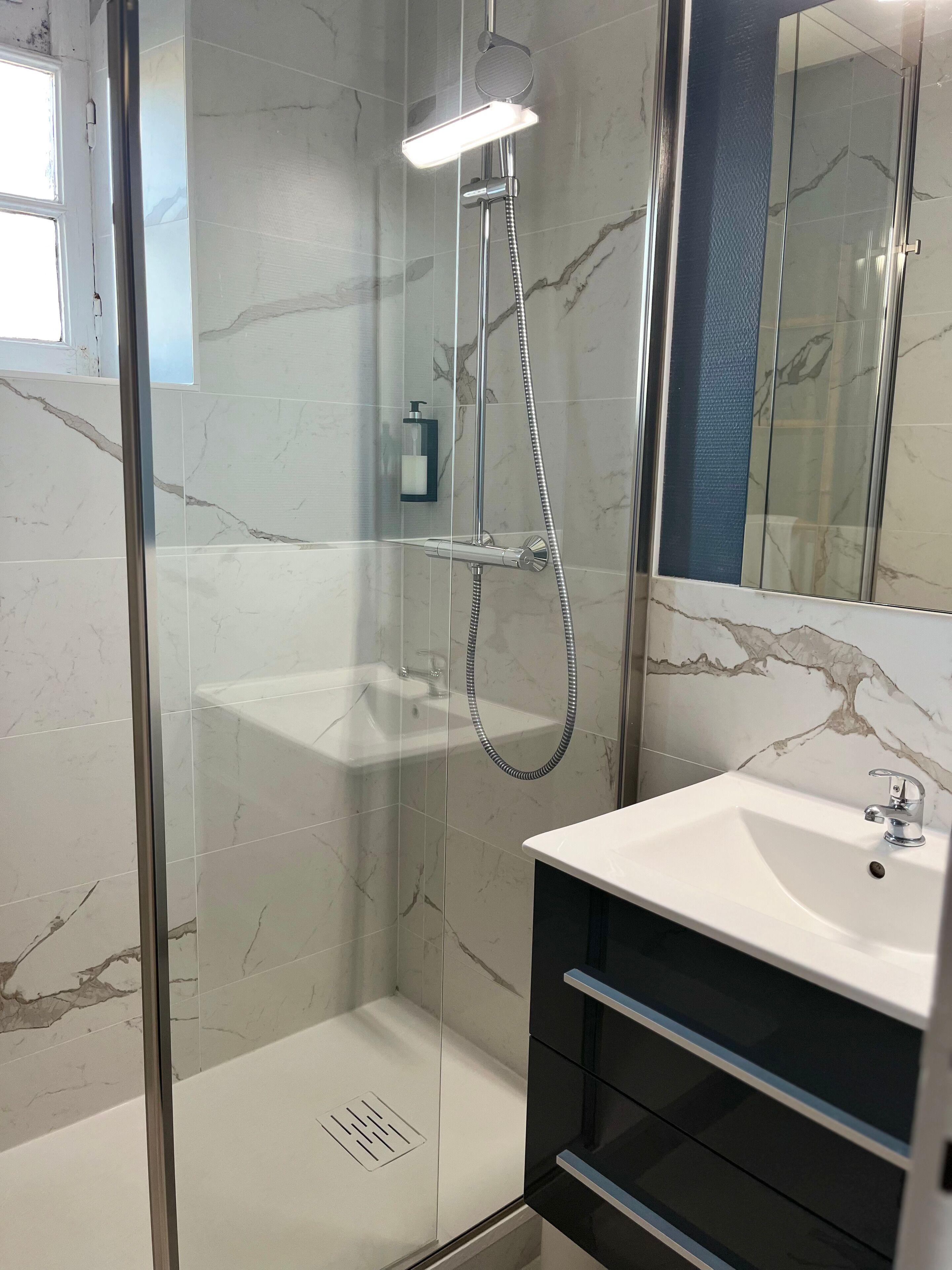 Un baño moderno con ducha de mampara de vidrio, lavamanos blanco y paredes con diseño de mármol.