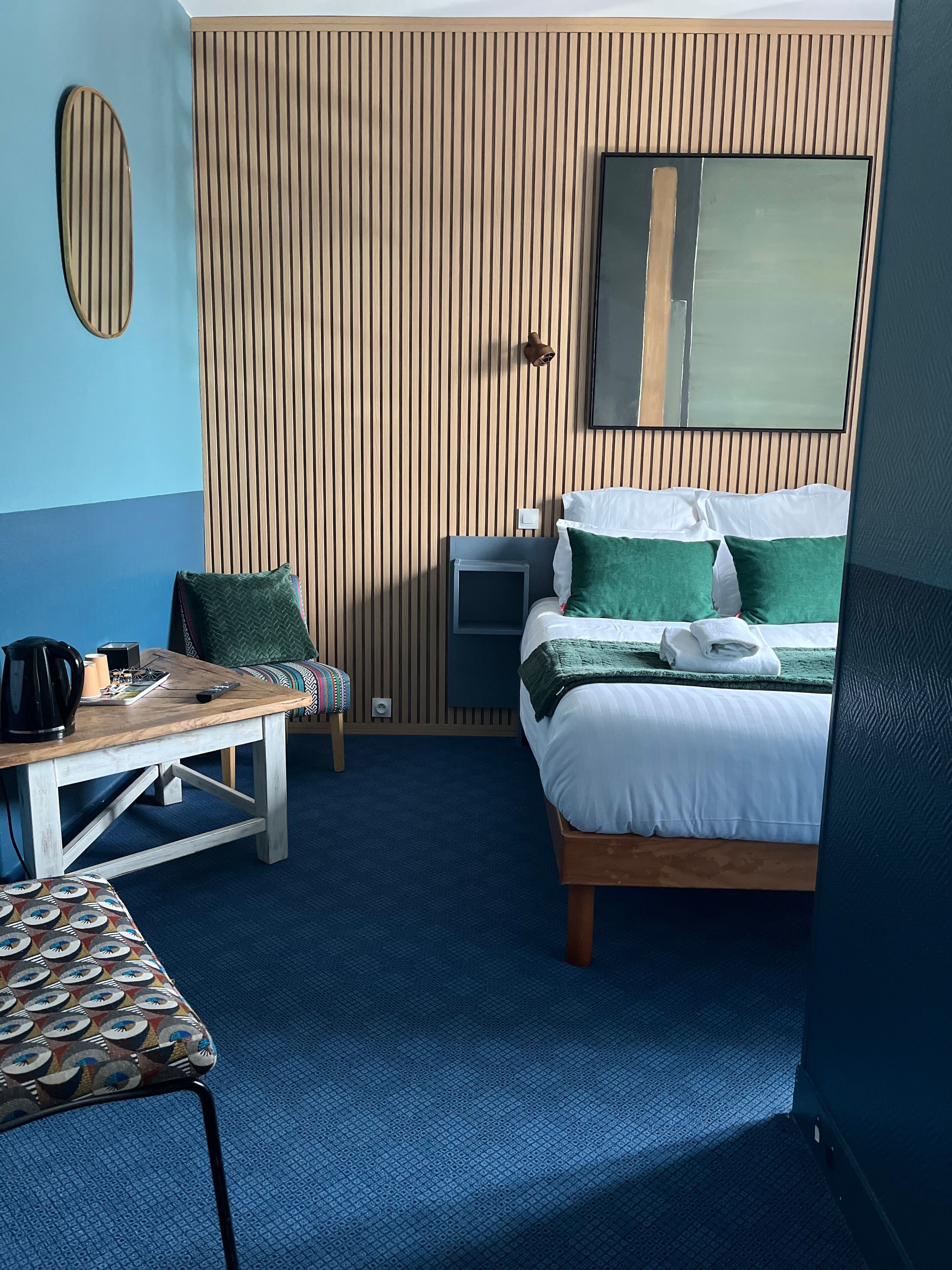 A hotel room with a bed, bedside table, chair, and a small desk with a lamp.
