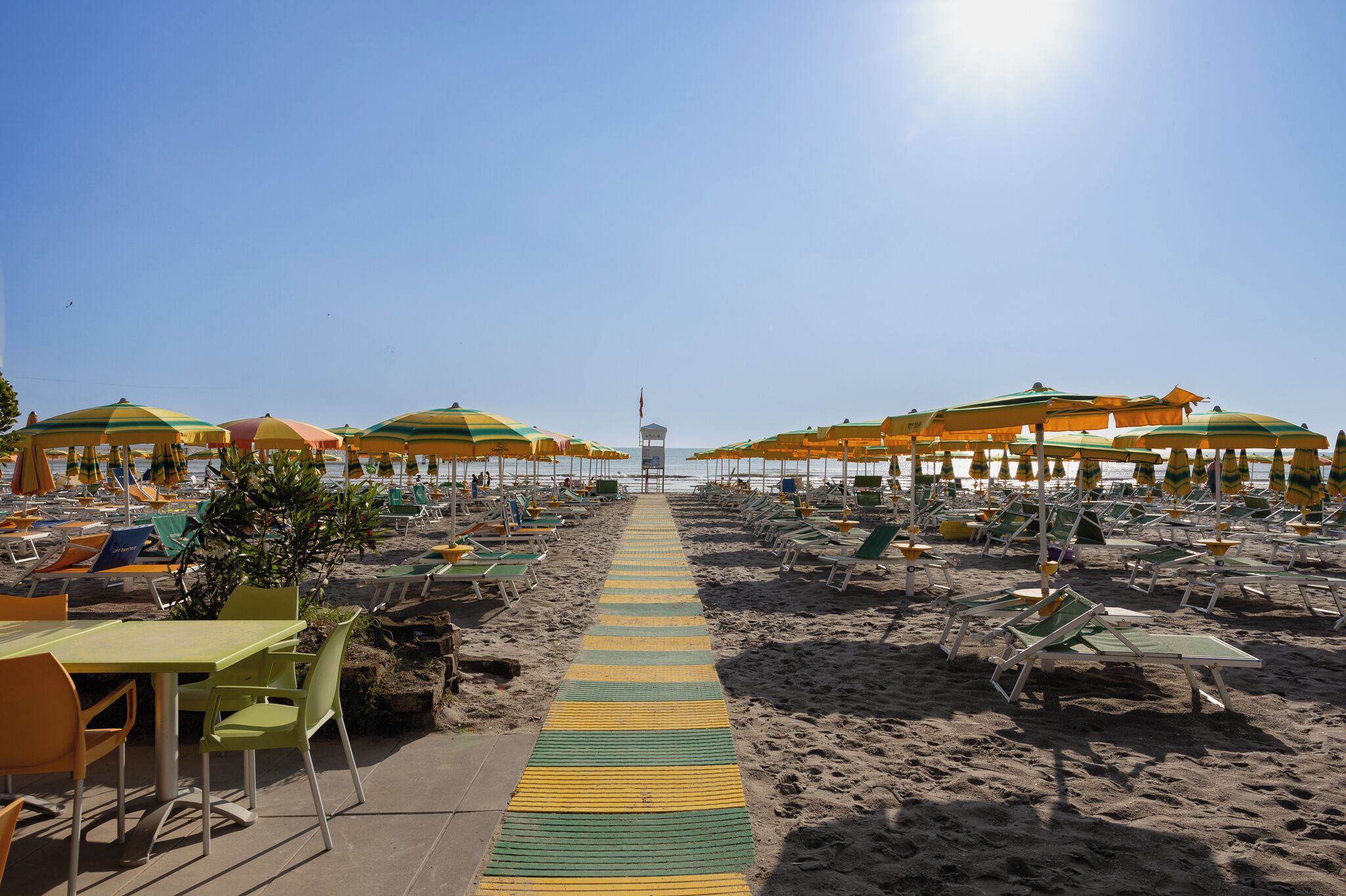 A beach with rows of sun loungers and umbrellas, a central walkway, and a view towards a distant structure.