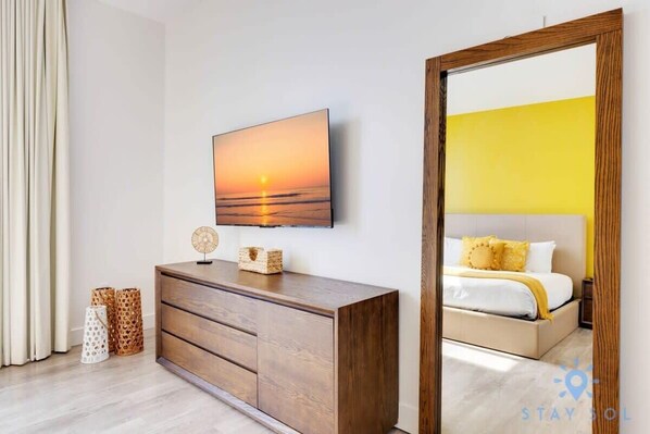 A modern bedroom with a wooden dresser, a flat-screen TV mounted on the wall, and a bed with yellow bedding.