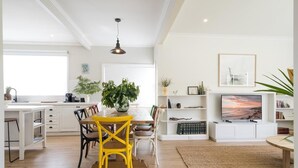 A modern kitchen with white cabinetry, a dining table with yellow chairs, a large potted plant, and a television in the living area. - Seafarers Cottage - A Dog's Dream on the Mornington Peninsula! (Capel Sound)