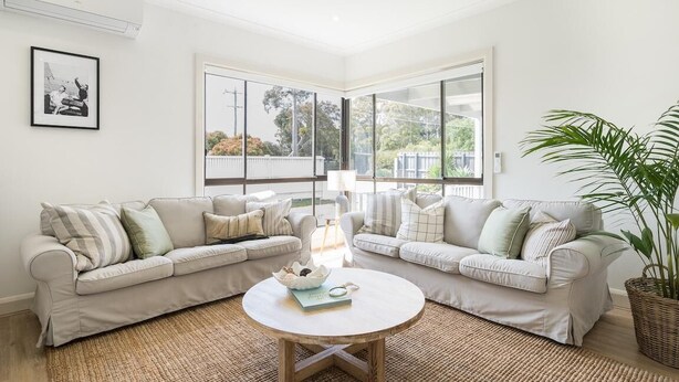 A modern living room with two beige sofas, a round coffee table, and a large window providing a view of the outdoors. - Seafarers Cottage - A Dog's Dream on the Mornington Peninsula! (Capel Sound)