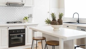A modern kitchen with white cabinetry, a central island with stools, a gas stove, and a hanging pendant light. - Seafarers Cottage - A Dog's Dream on the Mornington Peninsula! (Capel Sound)
