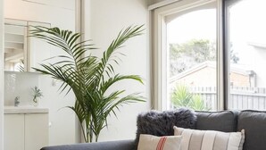 A modern living room with a grey sofa, a round wooden coffee table, and a large window offering a view of the outside. - Seafarers Cottage - A Dog's Dream on the Mornington Peninsula! (Capel Sound)