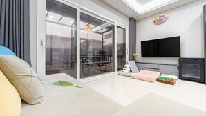 A modern living room with a sofa, a TV, a small table with a teddy bear, and a grill outside the sliding glass door. - Anmyeondo Town 1967 Dog Pool Villa (Taean)
