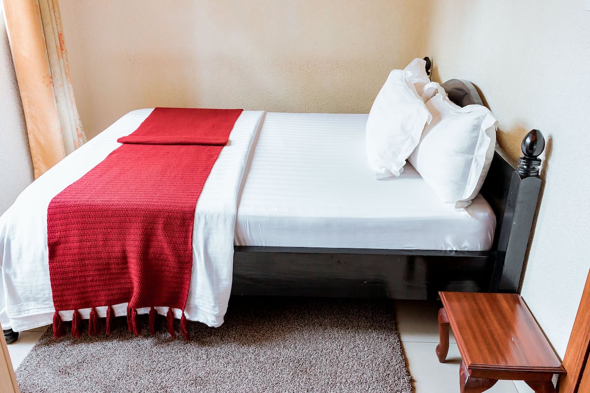 A neatly made bed with a red blanket and white pillows, a wooden nightstand, and a small rug on the floor.