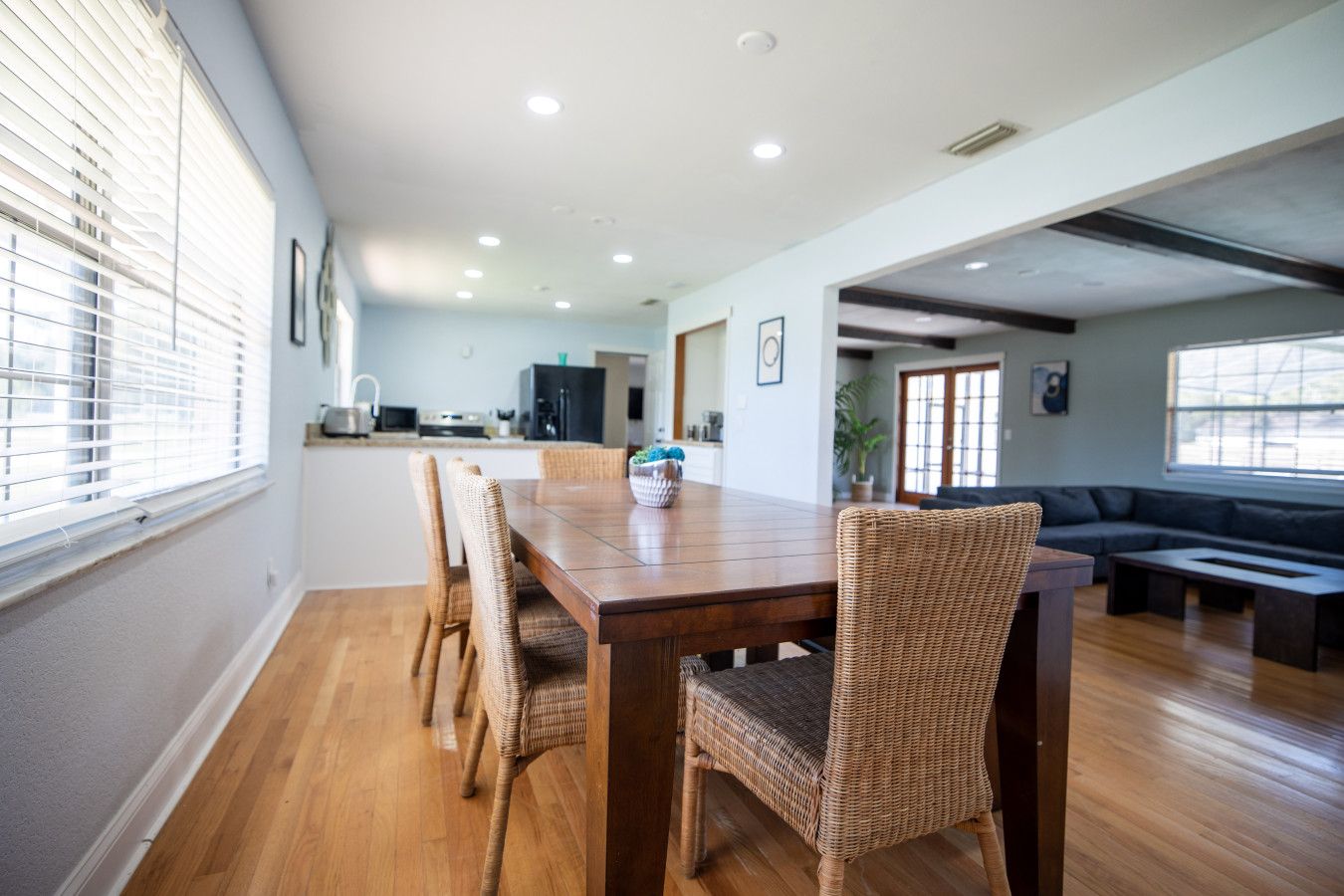 Un comedor amplio con una mesa de madera y sillas de mimbre, una cocina al fondo y un área de estar con un sofá y una mesita de centro.