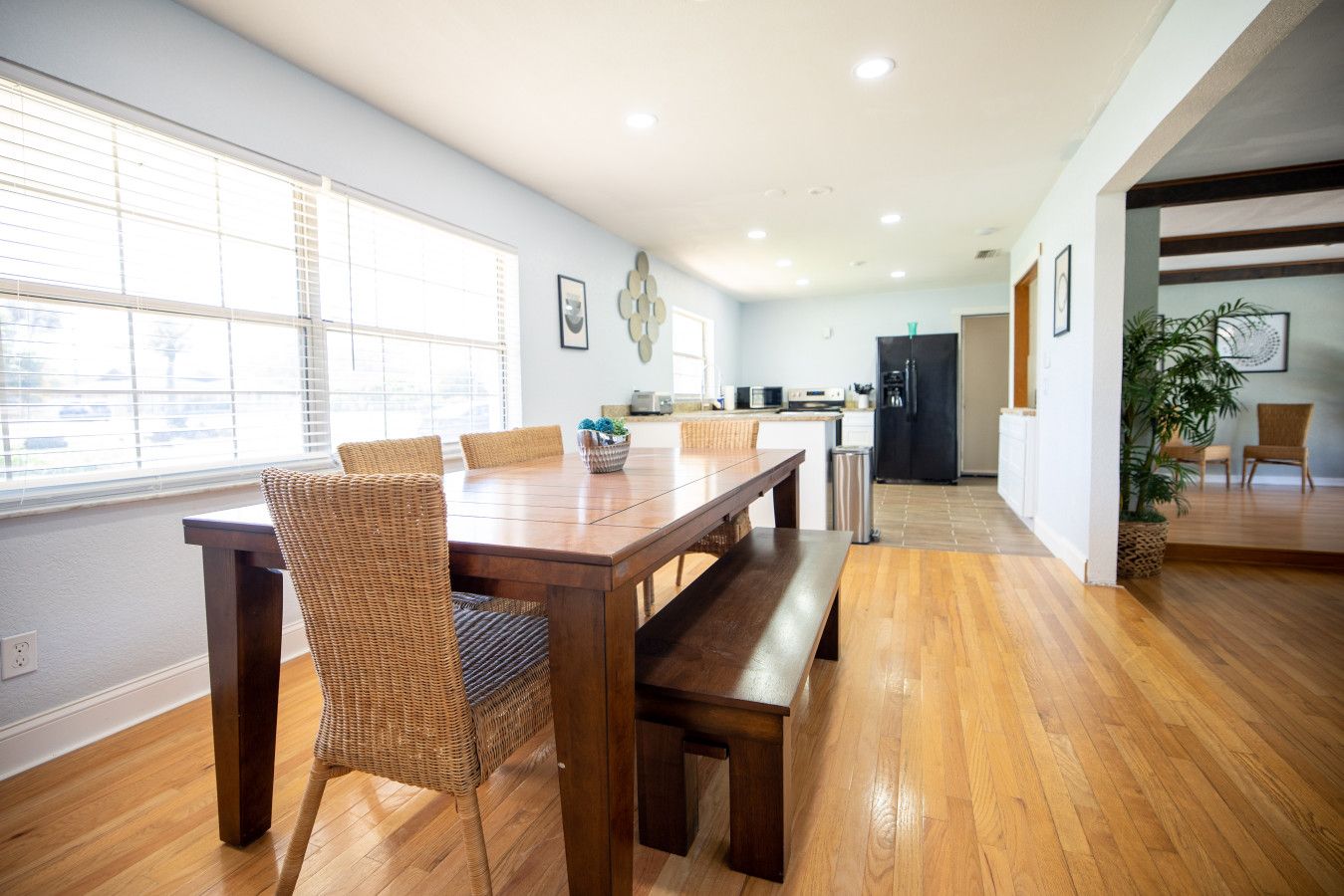 Uma sala de jantar espaçosa com mesa de madeira e cadeiras de vime, cozinha ao fundo e vista através de uma porta aberta.
