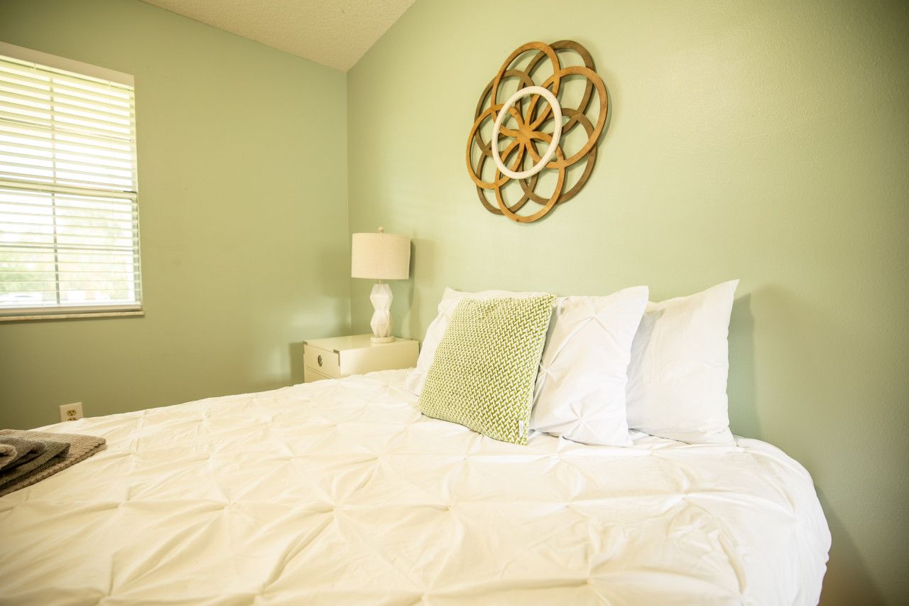 Cama bem arrumada com roupa de cama branca, um objeto decorativo na parede e uma luminária de cabeceira.